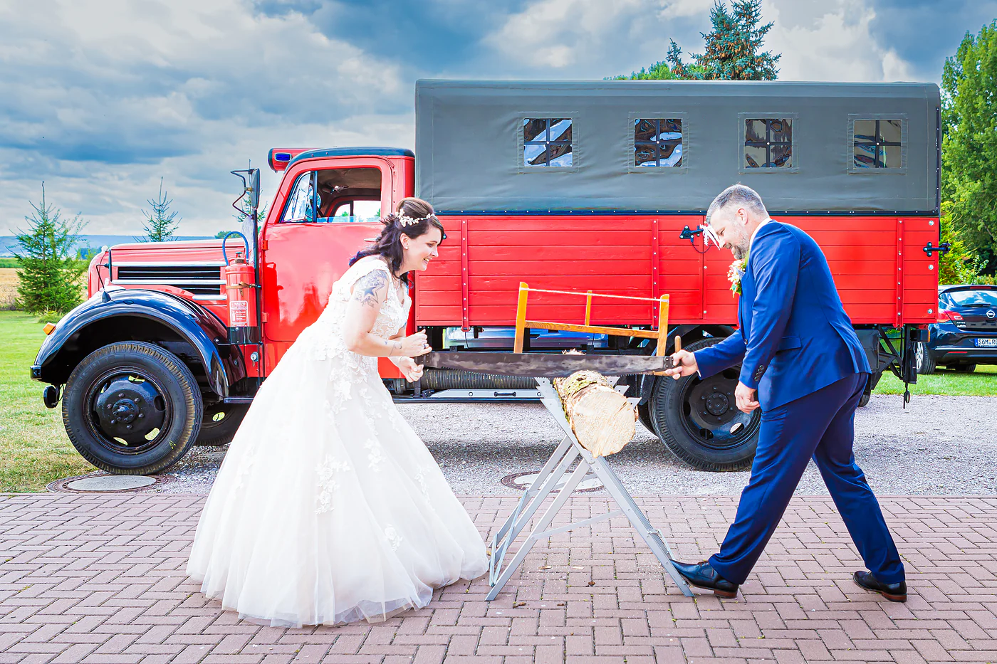 Brautpaar sägt vor dem roten Feuerwehr-Oldtimer gemeinsam einen Baumstamm durch, Hochzeitsbrauch nach der Ankunft