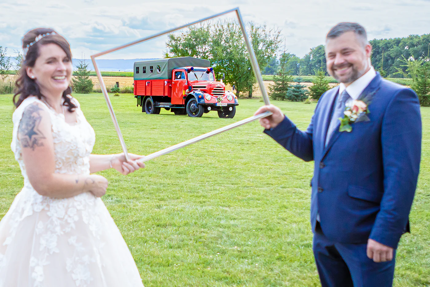 Brautpaar hält einen leeren Bilderrahmen hoch, darin steht der rote Feuerwehr-Oldtimer auf der Wiese