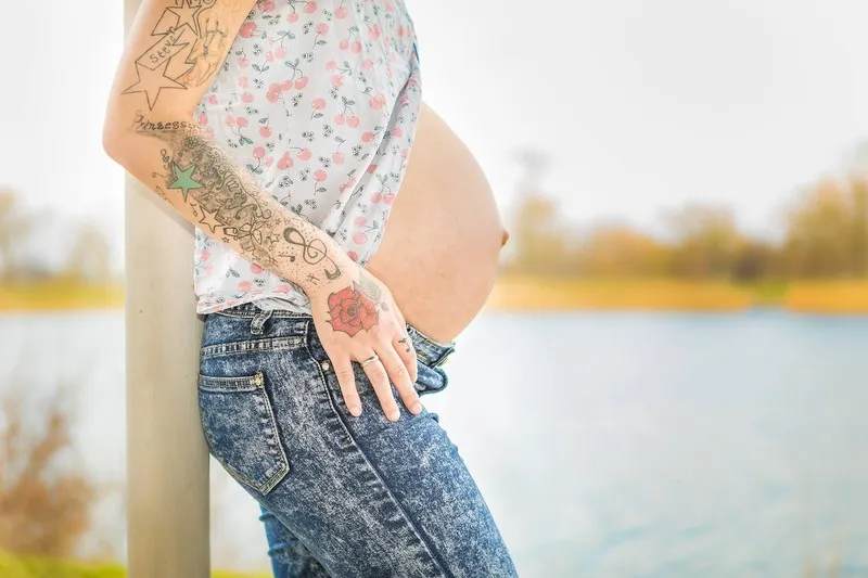 Seitliches Profil eines nackten Babybauchs am Wasser, die tätowierte Hand der werdenden Mutter ruht an der Jeans, Babybauch Shooting Sömmerda Dirk Dittrich