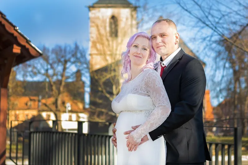 Schwangere Braut im weißen Spitzenkleid und Bräutigam im dunklen Anzug vor einem Kirchturm, seine Hände liegen auf ihrem Babybauch, Babybauch Shooting Sömmerda Dirk Dittrich
