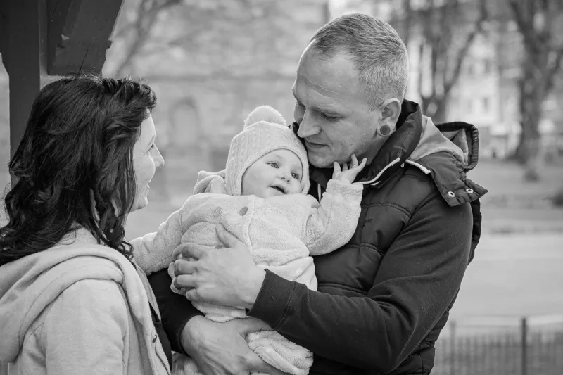 Schwarzweißaufnahme junger Eltern im Freien, der Vater hält das Baby mit Bommelmütze liebevoll im Arm, die Mutter schaut es lächelnd an, Babybauch Shooting Sömmerda Dirk Dittrich