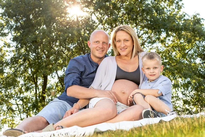 Werdende Eltern mit ihrem Sohn auf einer Picknickdecke in der Wiese, die Mutter zeigt ihren nackten Babybauch, dahinter Bäume im Gegenlicht, Babybauch Shooting Sömmerda Dirk Dittrich