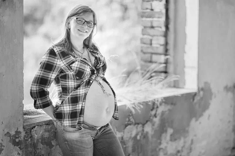 Schwarzweißaufnahme einer lächelnden Schwangeren mit Brille und offenem Karohemd, sie lehnt an einer alten Fensteröffnung mit Ziegelmauer, Babybauch Shooting Sömmerda Dirk Dittrich
