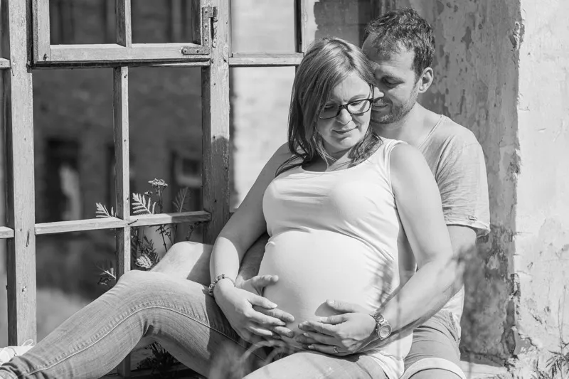 Schwarzweißaufnahme eines Paares an einem alten Sprossenfenster, der Mann sitzt hinter der Schwangeren und beide legen die Hände auf den Babybauch, Babybauch Shooting Sömmerda Dirk Dittrich