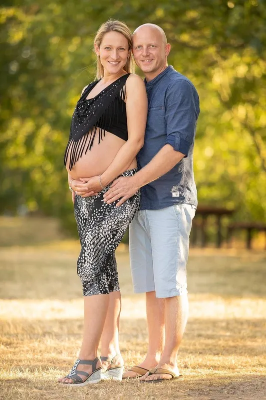 Schwangere Frau mit freiem Babybauch und ihr Partner stehen eng umschlungen im warmen Abendlicht eines Parks, Babybauch Shooting Sömmerda Dirk Dittrich