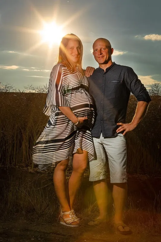 Werdende Eltern stehen bei tiefstehender Sonne am Feldrand, die Frau im gestreiften Kleid hält ihren Babybauch, dramatischer Sonnenstern am Himmel, Babybauch Shooting Sömmerda Dirk Dittrich