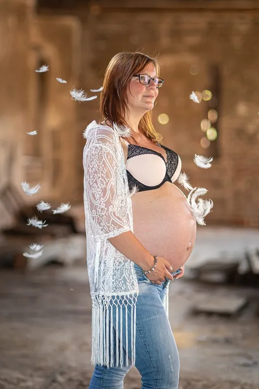 Schwangere in weißem Spitzenkimono steht in einem alten Gebäude, um sie herum schweben weiße Federn, Babybauch Shooting Sömmerda Dirk Dittrich