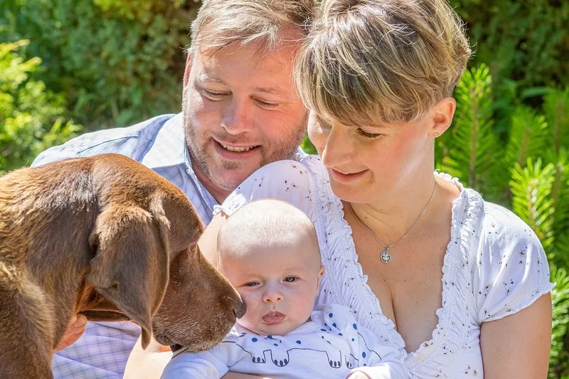 Eltern halten ihr Baby, während der braune Familienhund neugierig daran schnuppert, sommerliches Grün im Hintergrund, Familienfotograf Sömmerda Dirk Dittrich