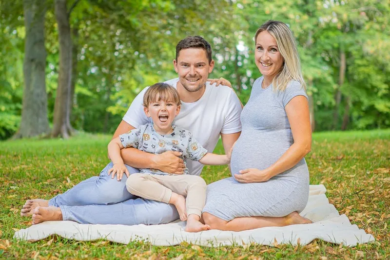 Schwangere Frau mit Babybauch sitzt mit Partner und lachendem Kleinkind auf einer Decke im grünen Park, Familienfotograf Sömmerda Dirk Dittrich