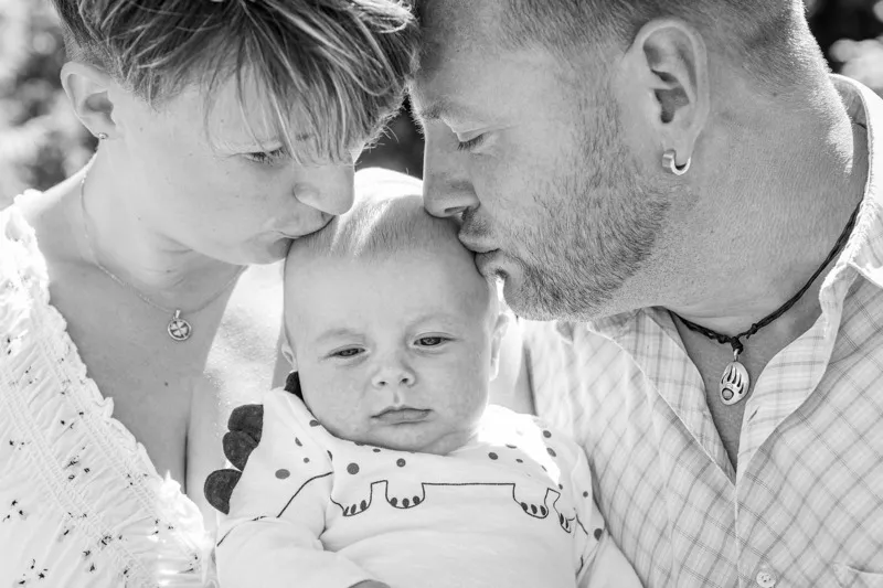 Schwarzweiß-Aufnahme: Mutter und Vater küssen ihr Baby gleichzeitig zärtlich auf den Kopf, Familienfotograf Sömmerda Dirk Dittrich
