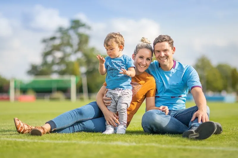 Eltern sitzen mit ihrem Kleinkind auf dem Rasen eines Sportplatzes, im Hintergrund ein Fußballtor unter blauem Himmel, Familienfotograf Sömmerda Dirk Dittrich