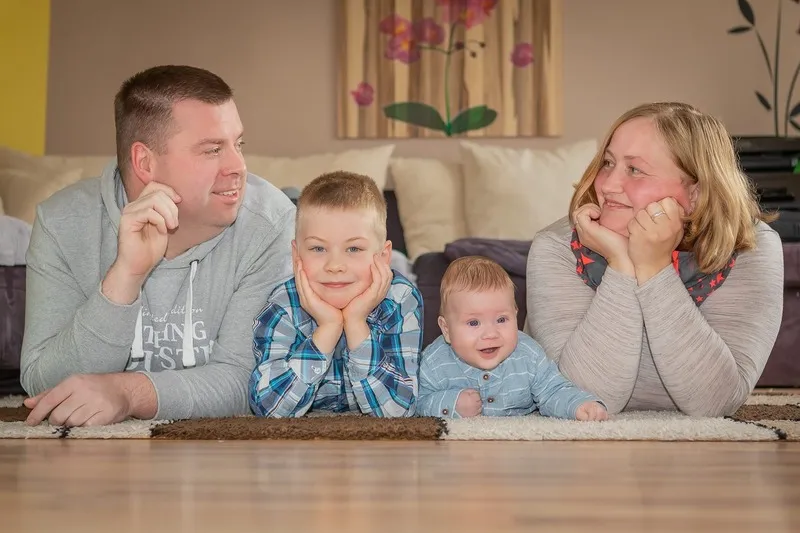 Familie mit zwei Kindern liegt auf dem Teppich im Wohnzimmer, der große Bruder stützt das Kinn auf, das Baby strahlt, Familienfotograf Sömmerda Dirk Dittrich