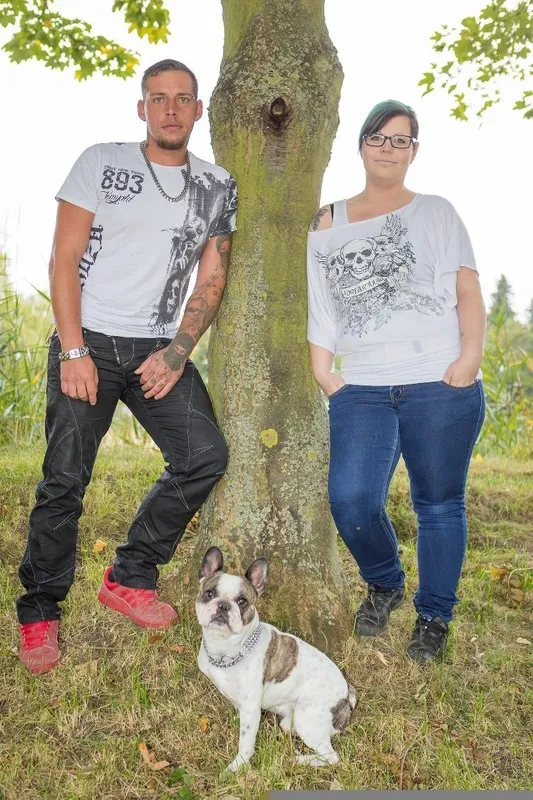 Paar steht lässig an einem Baumstamm, davor sitzt ihre Französische Bulldogge im Gras, Familienfotograf Sömmerda Dirk Dittrich