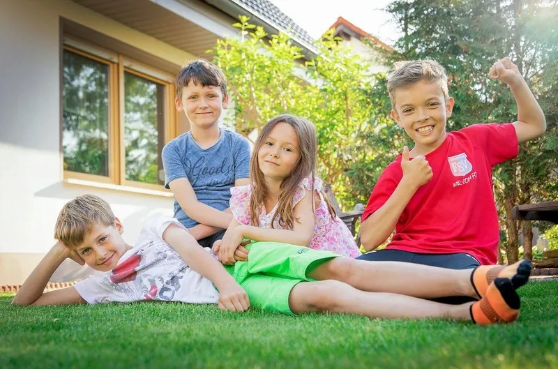 Vier fröhliche Kinder sitzen und liegen auf dem grünen Rasen vor einem Haus, ein Junge jubelt mit erhobener Faust, Familienfotograf Sömmerda Dirk Dittrich