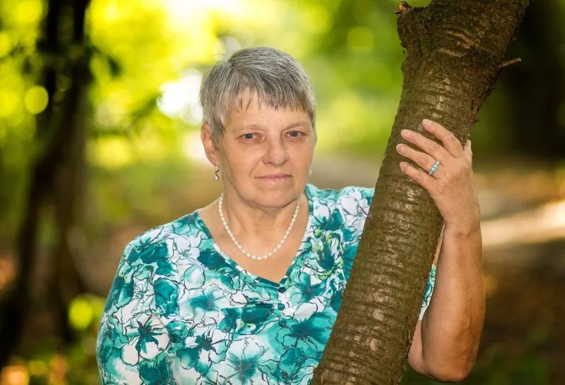 Frau mit Perlenkette und geblümter Bluse lehnt an einem Baumstamm im sonnigen Wald, Portraitfotograf Sömmerda Dirk Dittrich