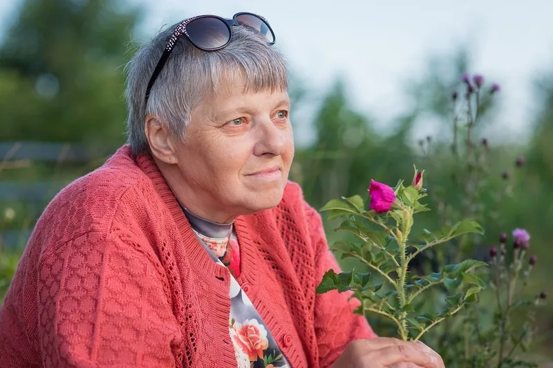 Frau in korallenroter Strickjacke mit Sonnenbrille im Haar neben einer blühenden Rose im sommerlichen Garten, Portraitfotograf Sömmerda Dirk Dittrich