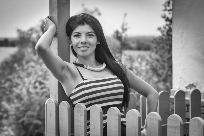 Schwarzweiß Portrait einer lächelnden Frau mit langen dunklen Haaren im gestreiften Kleid an einem Holzzaun, Portraitfotograf Sömmerda Dirk Dittrich