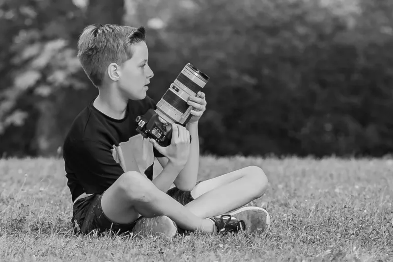 Junge sitzt im Schneidersitz auf der Wiese und hält eine Spiegelreflexkamera mit Teleobjektiv, Schwarzweiß Aufnahme, Portraitfotograf Sömmerda Dirk Dittrich