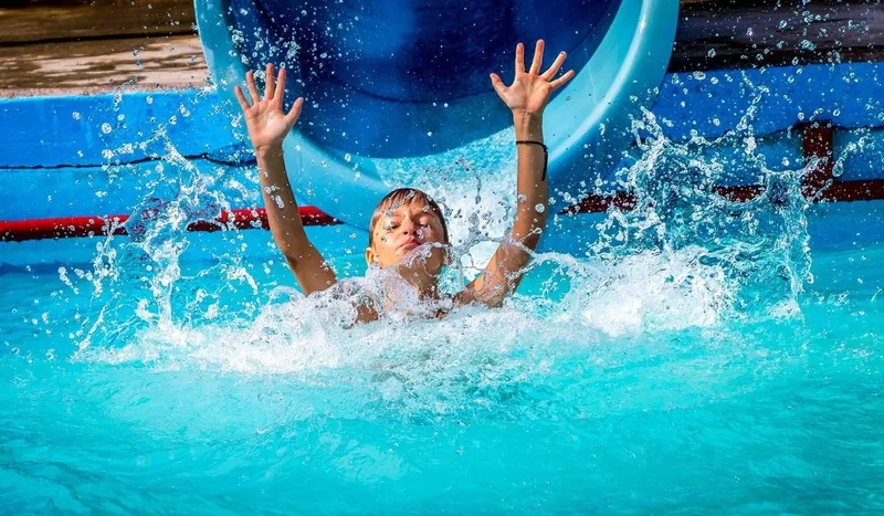 Junge landet mit hochgerissenen Armen und spritzendem Wasser am Ende einer blauen Wasserrutsche im Pool, Portraitfotograf Sömmerda Dirk Dittrich