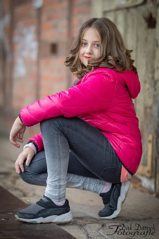 Mädchen in pinker Steppjacke hockt vor einer alten Backsteinmauer und blickt über die Schulter, Portraitfotograf Sömmerda Dirk Dittrich