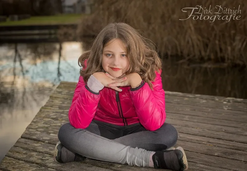 Mädchen in pinker Jacke sitzt im Schneidersitz auf einem Holzsteg am Teich, Portraitfotograf Sömmerda Dirk Dittrich