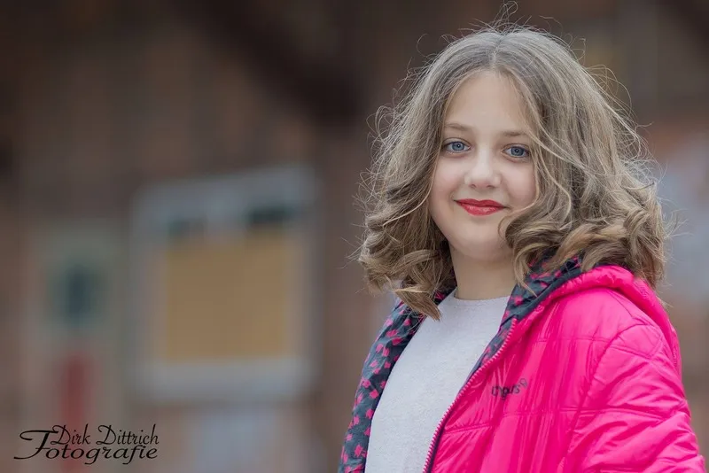 Lächelndes Mädchen mit gelockten Haaren in pinker Steppjacke vor unscharfer Backsteinfassade, Portraitfotograf Sömmerda Dirk Dittrich