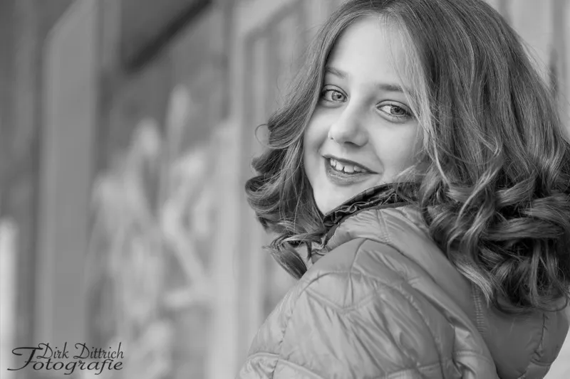 Schwarzweiß Portrait eines lächelnden Mädchens mit Locken, das in Steppjacke über die Schulter blickt, Portraitfotograf Sömmerda Dirk Dittrich
