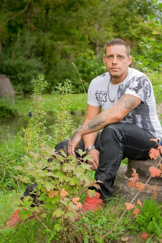 Junger Mann mit Tätowierungen sitzt nachdenklich auf einem Stein am Bachufer im Grünen, Portraitfotograf Sömmerda Dirk Dittrich