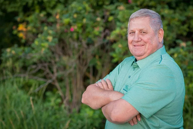 Freundlich lächelnder Mann mit grauem Haar und verschränkten Armen in grünem Poloshirt vor sommerlichen Sträuchern, Portraitfotograf Sömmerda Dirk Dittrich